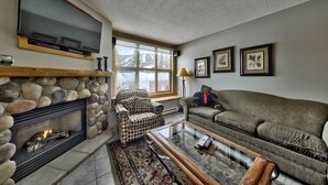 A cozy living room with a stone fireplace, a large flat-screen TV, a patterned armchair, a sofa, and a coffee table.