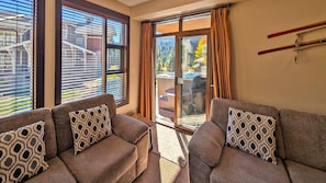 A room with two sofas, patterned cushions, a sliding glass door, and a view of a mountain landscape. - Stones Throw #12 (Sun Peaks)