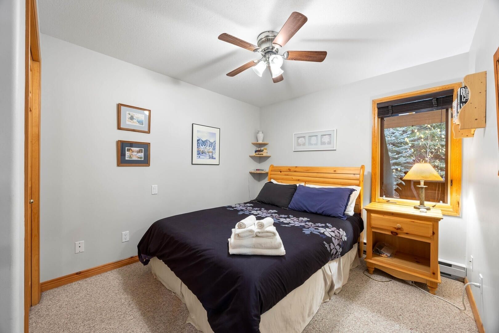 A bedroom with a bed, a wooden headboard, a ceiling fan, a nightstand with a lamp, and framed pictures on the wall.