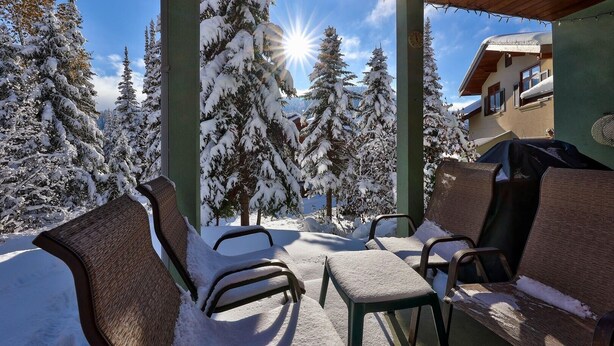 Un espacio de estar al aire libre techado con sillas y mesas cubiertas de nieve, con vistas a un paisaje nevado con árboles y un edificio al fondo.
