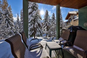 Ein überdachter Außenbereich mit schneebedeckten Stühlen und Tischen, mit Blick auf eine verschneite Landschaft mit Bäumen und einem Gebäude im Hintergrund.