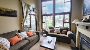 A living room with two sofas, a coffee table, a fireplace, and large windows with curtains. - Stones Throw #38 By Bear Country (Sun Peaks)
