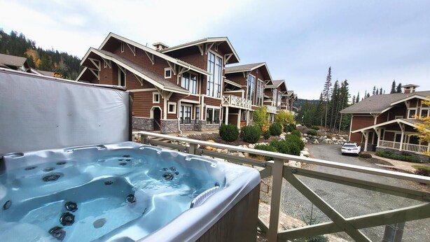A hot tub on a deck with a view of a large wooden building with multiple windows and balconies. - Stones Throw #38 By Bear Country (Sun Peaks)