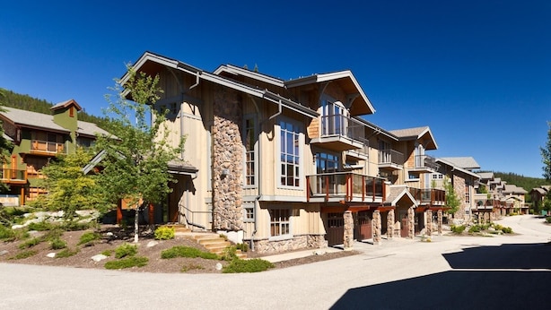 A row of multi-story wooden houses with stone accents, balconies, and a clear blue sky. - Trappers Landing #27 (Sun Peaks)