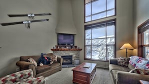 A cozy living room with a fireplace, large windows with a snowy view, and a bear statue on the couch. - Trappers Landing #27 (Sun Peaks)