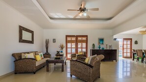 A spacious living room with a ceiling fan, a dining area with wicker furniture, and a large window with a view. - Grand home with a private pool, close to beach (Noord)
