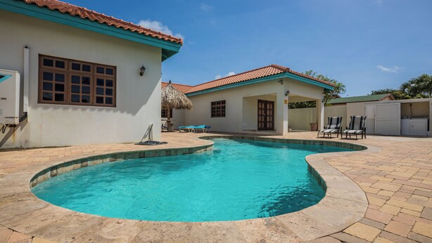 A swimming pool with a curved edge, surrounded by a patio area with tiled flooring and lounge chairs. - Grand home with a private pool, close to beach (Noord)