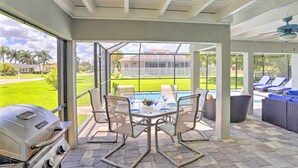 A covered patio with a grill, dining table, and chairs. There is a view of a swimming pool and a house in the background.