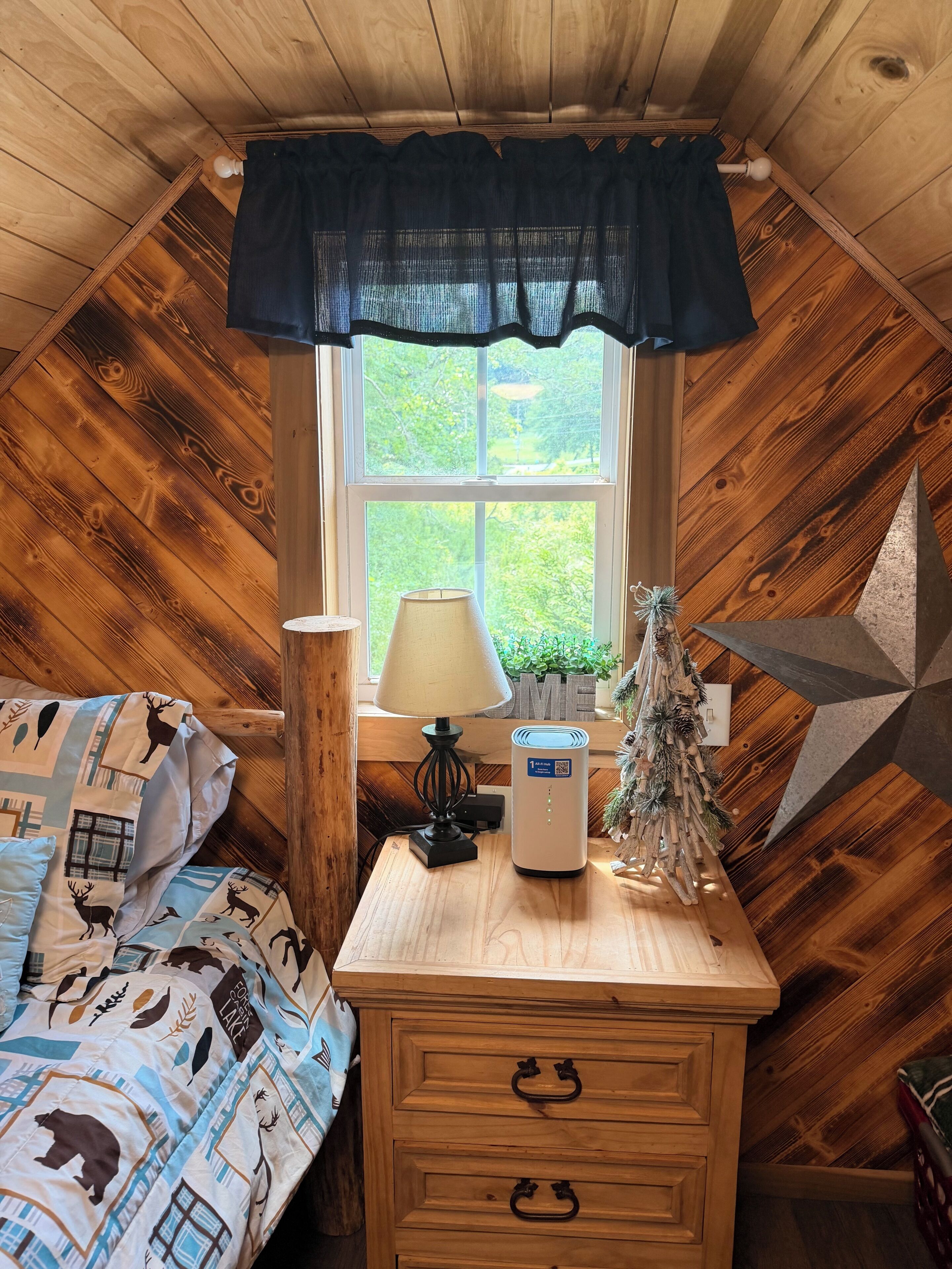 A cozy room with a sloped ceiling, a bed with a patterned blanket, a bedside table with a lamp, a decorative tree, and a wall-mounted shelf.