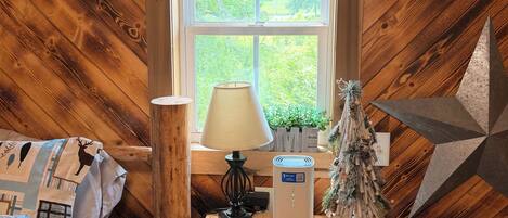 A cozy room with a sloped ceiling, a bed with a patterned blanket, a bedside table with a lamp, a decorative tree, and a wall-mounted shelf.