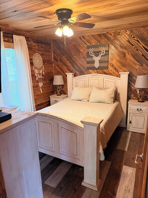 Une chambre avec un plafond en bois, un lit avec une literie blanche, deux tables de chevet avec des lampes, un attrape-rêves accroché au mur et un plancher en bois.
