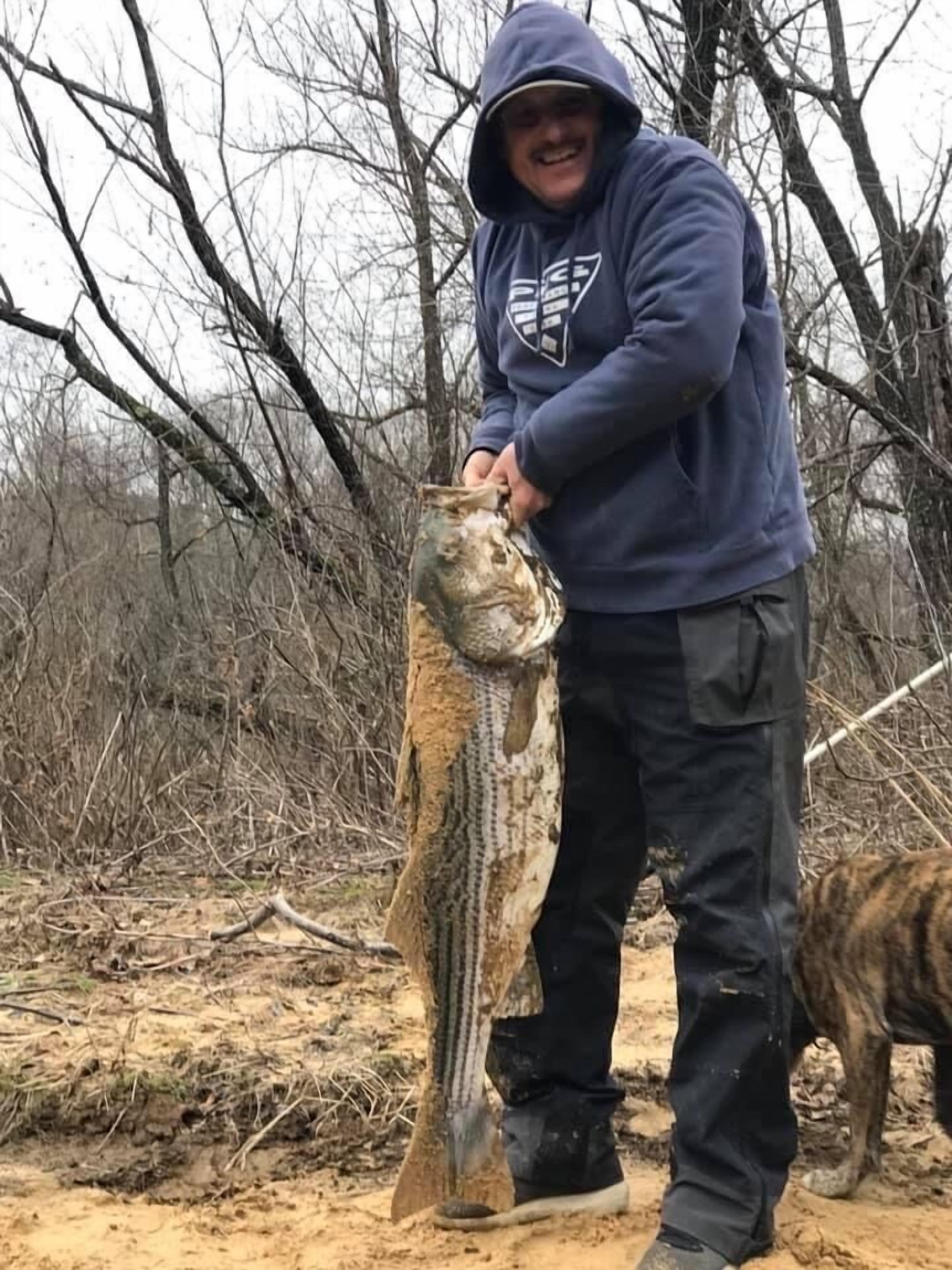 A person holding a fish in a natural outdoor setting with trees and a dog nearby.