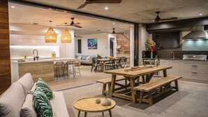 A modern kitchen with a central island, dining area with wooden tables and benches, and a cozy seating area with a sofa and a small round table. - Azure Port Douglas - Luxurious & Modern Villa (Port Douglas)