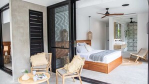 A modern bedroom with a large bed, a sitting area with chairs, a small table, and a bathroom with a patterned shower. - Azure Port Douglas - Luxurious & Modern Villa (Port Douglas)