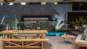 An outdoor dining area with a pool, wooden tables, and a stone wall backdrop. - Azure Port Douglas - Luxurious & Modern Villa (Port Douglas)