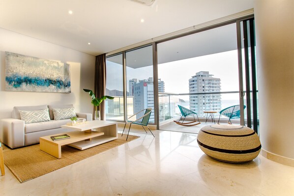 A modern living room with a sofa, coffee table, and ottoman, large windows with a city view, and a painting on the wall.