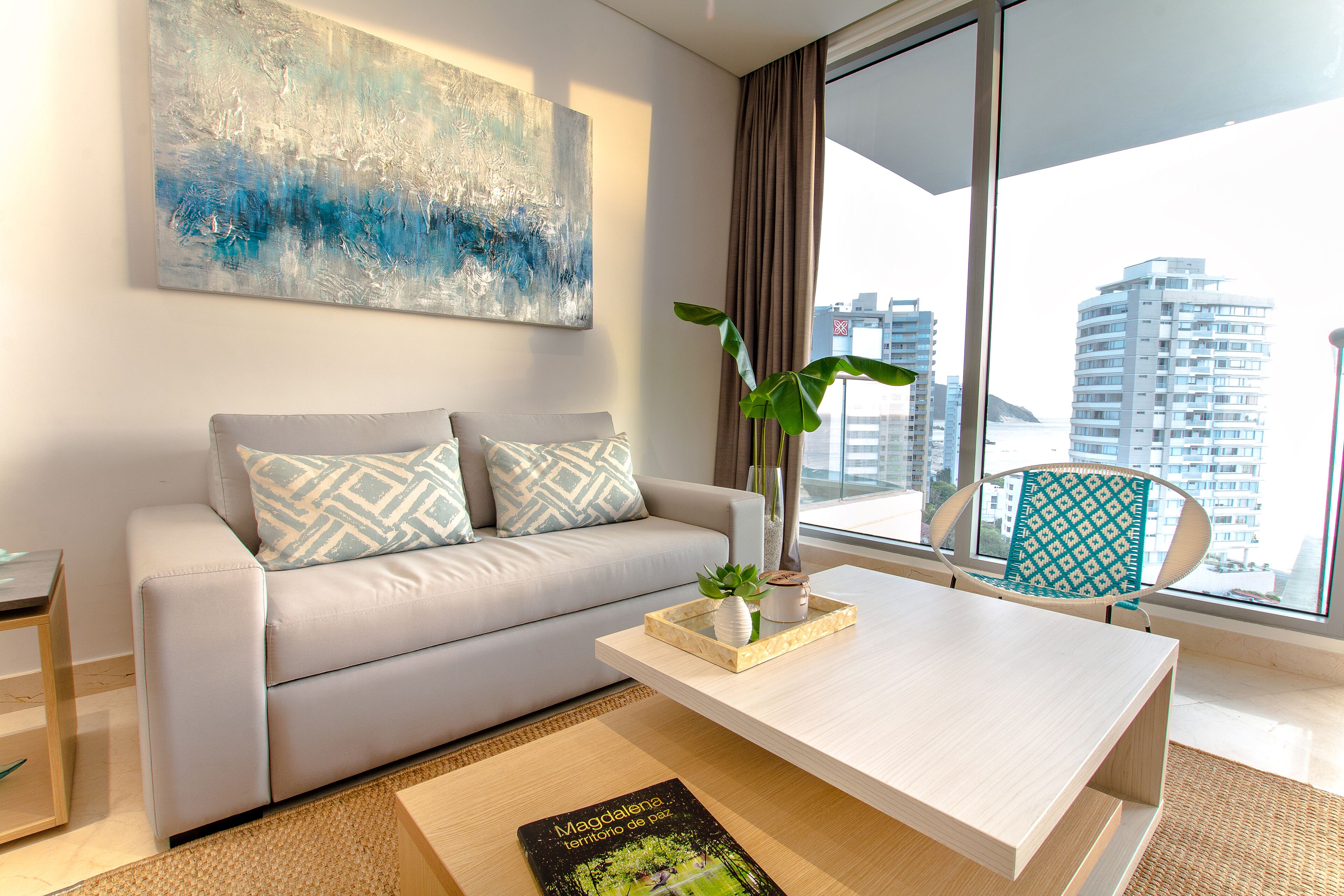 A modern living room with a sofa, a coffee table, and a view of the cityscape through large windows.