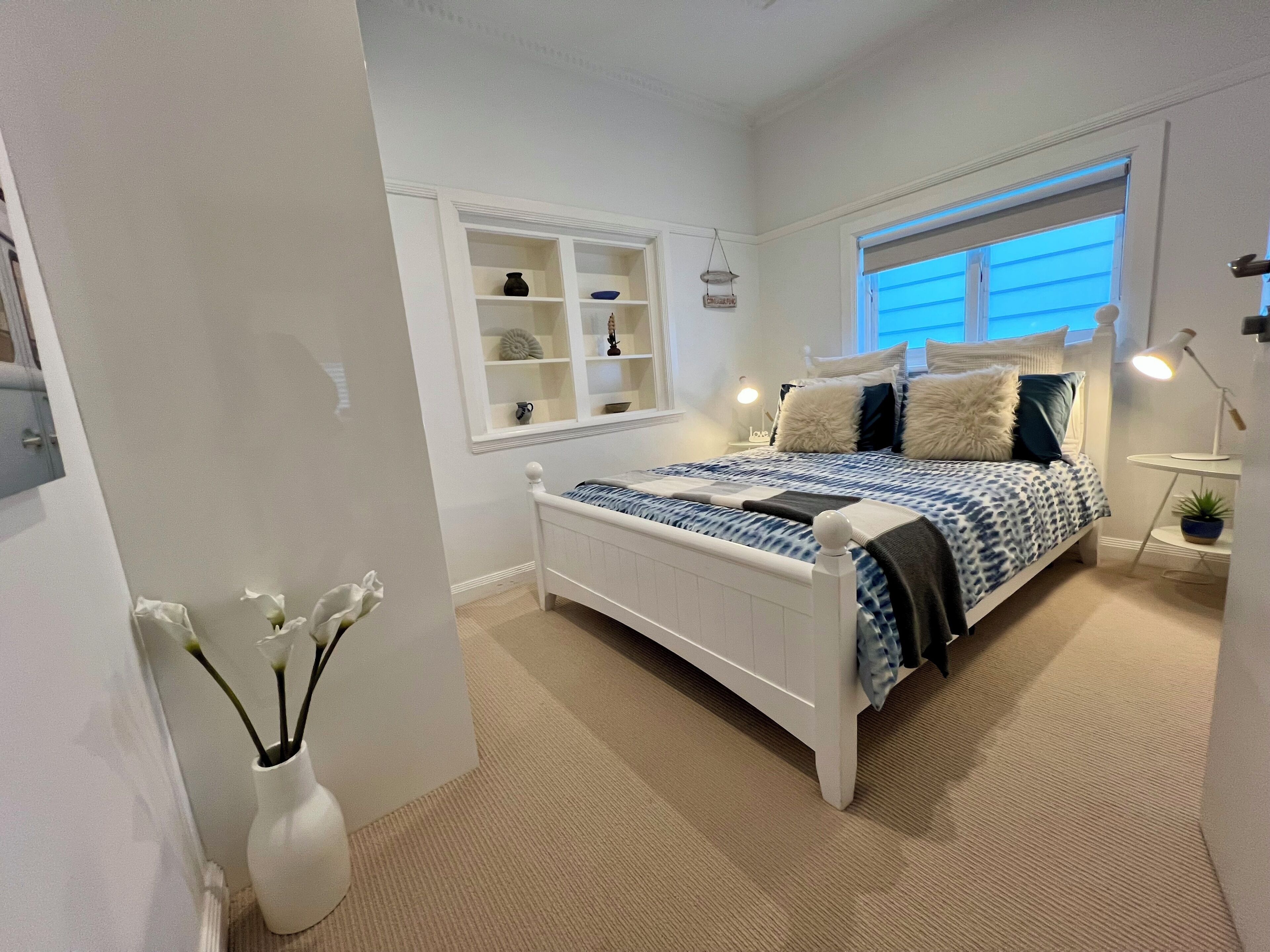 A bedroom with a white bed, a bedside table with a lamp, a window with blinds, and a white shelf unit.