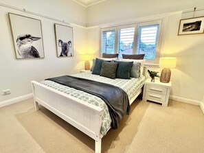 A bedroom with a bed, bedside lamps, a window with blinds, and framed pictures on the wall.