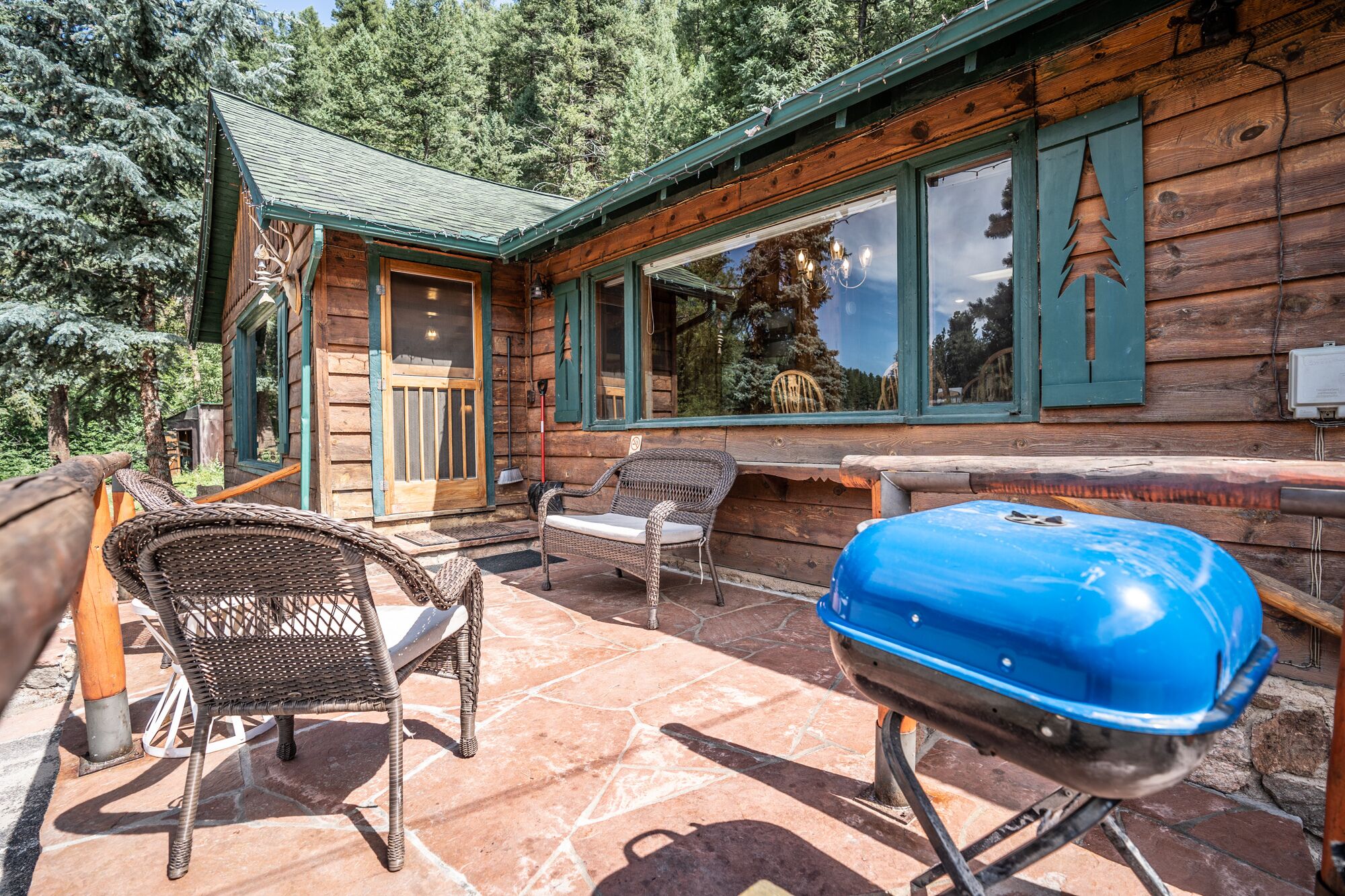 Una baita di legno con un patio in pietra, sedie in vimini e un barbecue blu.