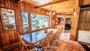 Comedor de madera con una gran mesa y sillas, una araña y vistas al exterior.