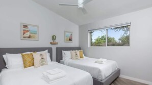 A hotel room with two beds, a ceiling fan, a large window, and artwork on the walls.