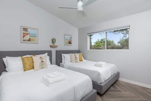 A hotel room with two beds, a ceiling fan, a large window, and artwork on the walls.