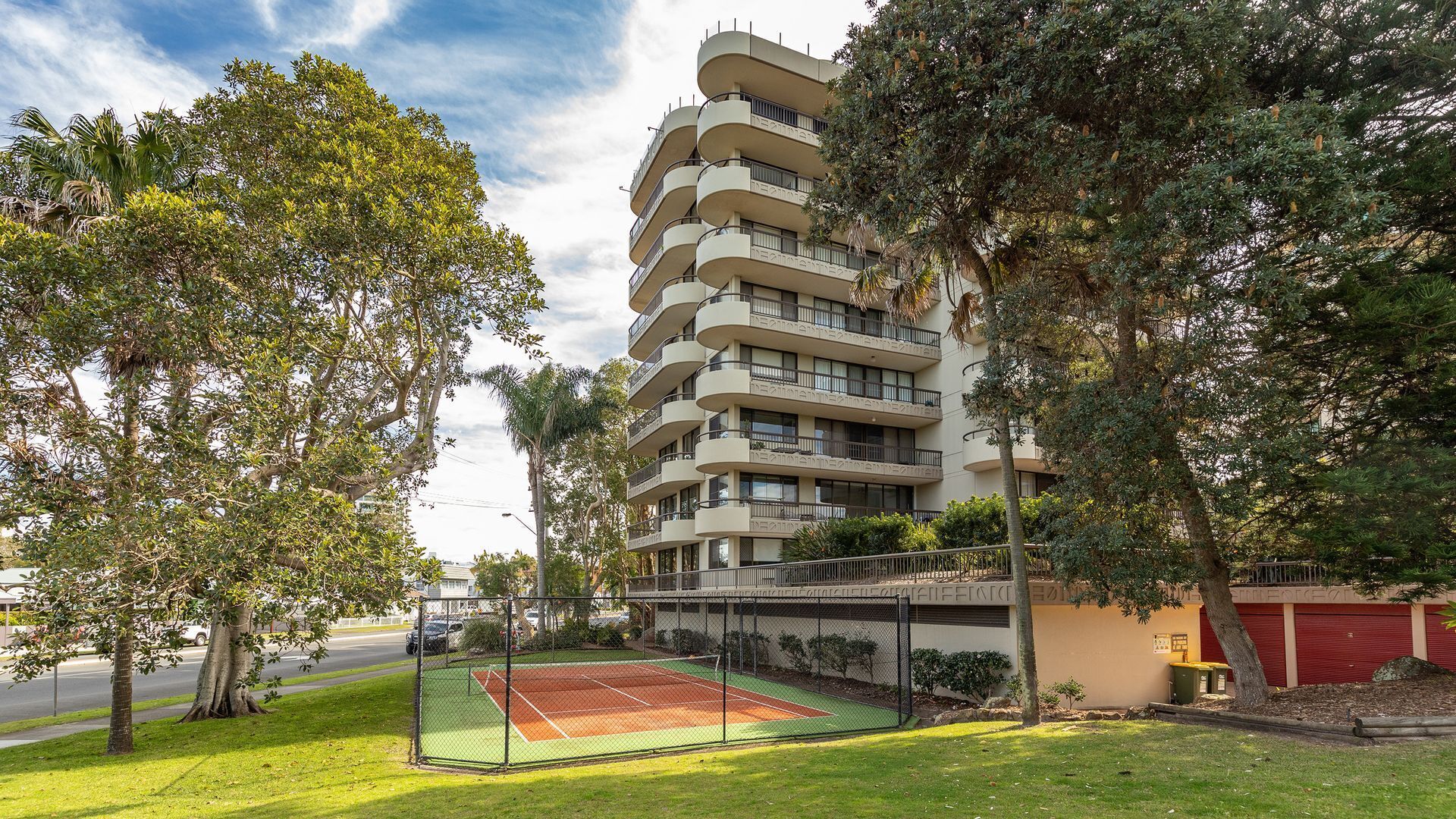 Un edificio residencial de varios pisos con balcones, rodeado de árboles y una cancha de tenis.