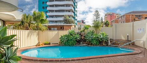 A swimming pool with a brick border, surrounded by plants and a fence.