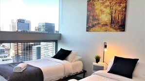 A hotel room with two beds, a city view, a painting of a forest, and a small plant on a bedside table.