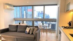 A modern living room with a grey sofa, a small round table, and a view of a cityscape through large windows.