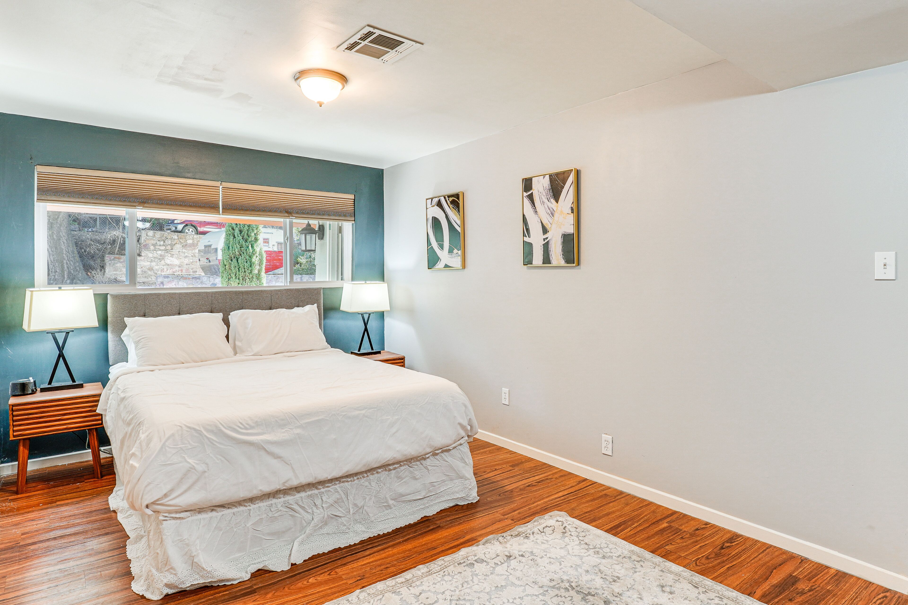 A bedroom with a large bed, two bedside tables with lamps, a window with blinds, and two abstract wall art pieces.