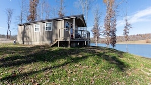 Eine Holzhütte mit Veranda, in der Nähe von Wasser gelegen und von Bäumen umgeben.