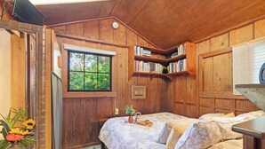A cozy cabin bedroom with wooden walls, a large window, a bookshelf, and a bed with a floral bedspread. - 1 Mile Away from Volcano National Park (Volcano)