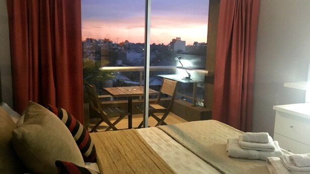 Ein Hotelzimmer mit Bett, einem kleinen Tisch und einem Stuhl. Durch ein großes Fenster bietet sich Blick auf die Stadt.