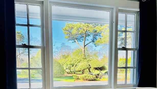 A room with a bay window, two white chairs, a small round table, and a view of a garden with trees and a lawn. - Sandy Neck Motel (East Sandwich)