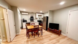 A compact kitchen with white cabinets, a stainless steel refrigerator, and a microwave. There is a wooden dining table with four chairs. A flat-screen TV is on a stand to the right. - Sandy Neck Motel (East Sandwich)