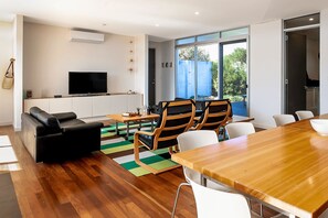 A modern living room with a flat-screen TV, a sofa, a dining table with chairs, and a large window offering a view of the outdoors.