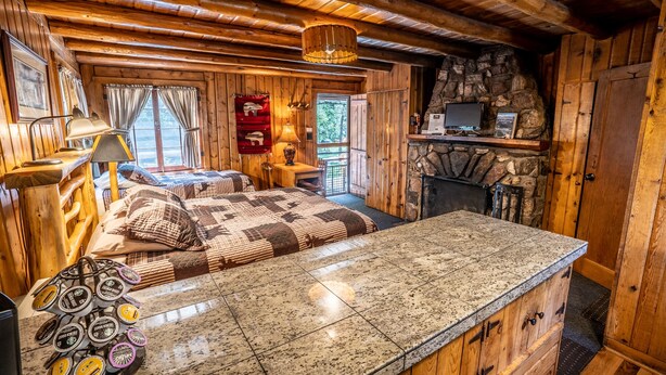Ein gemütliches Blockhaus-Interieur mit Holzbalkendecke, Kamin aus Stein, einem Bett mit karierter Decke und einer Frühstücksterrasse mit Kaffeetassen.