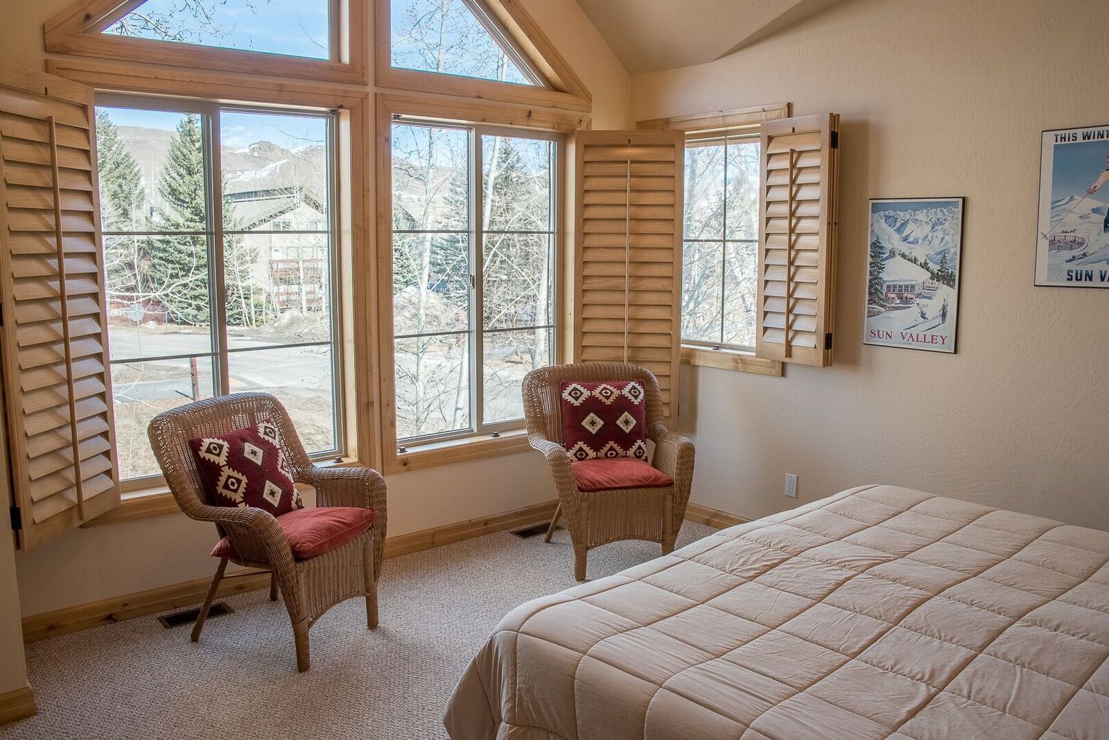 Una habitación con dos sillones de mimbre con cojines rojos, una cama grande y un cartel con una escena de montaña nevada.