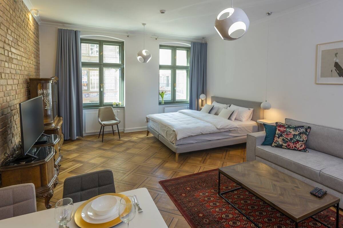 A modern hotel room with a bed, a sofa, a dining table with chairs, a TV, and a fireplace.