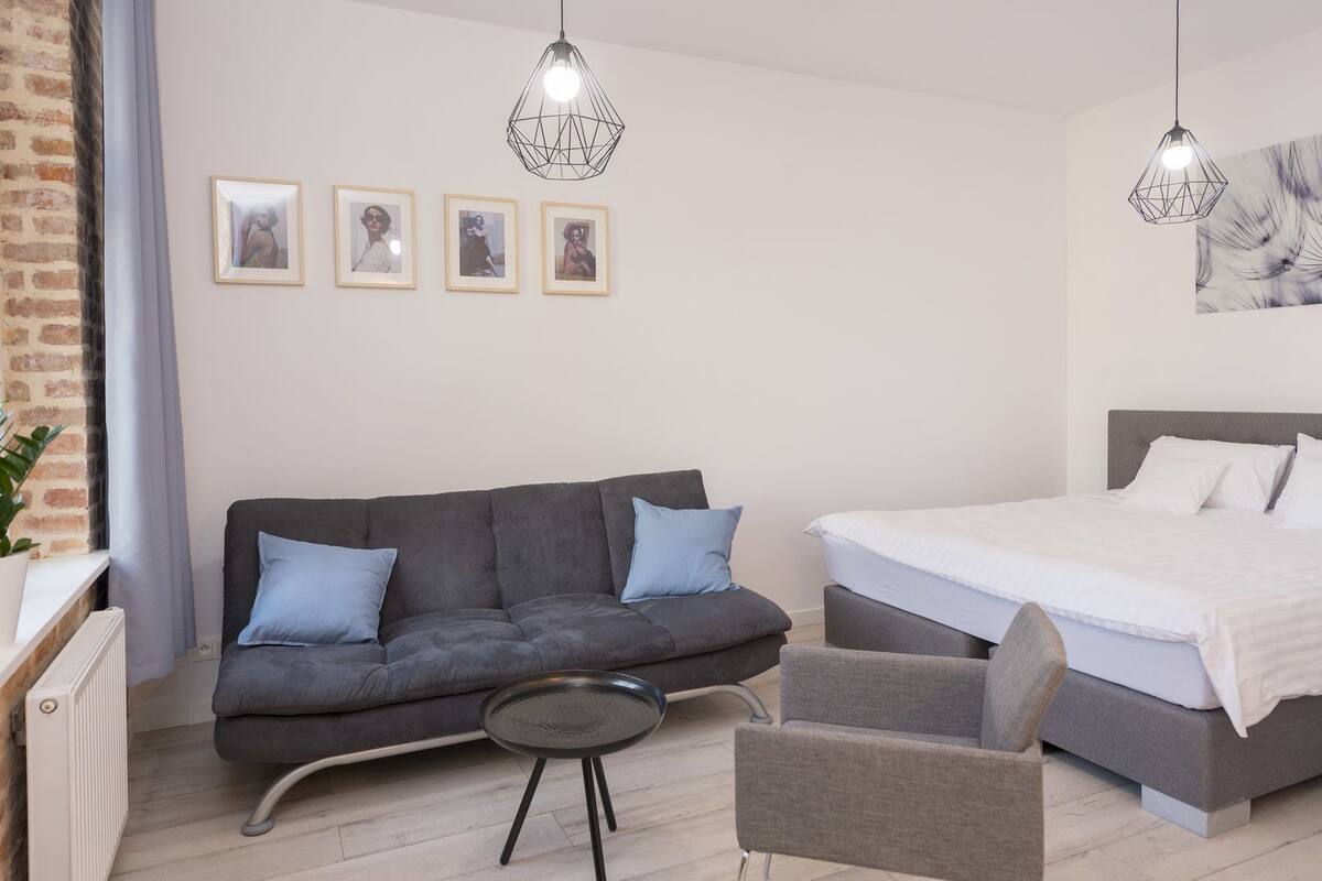 A modern bedroom with a grey sofa, a small round table, a single bed, and two pendant lights.
