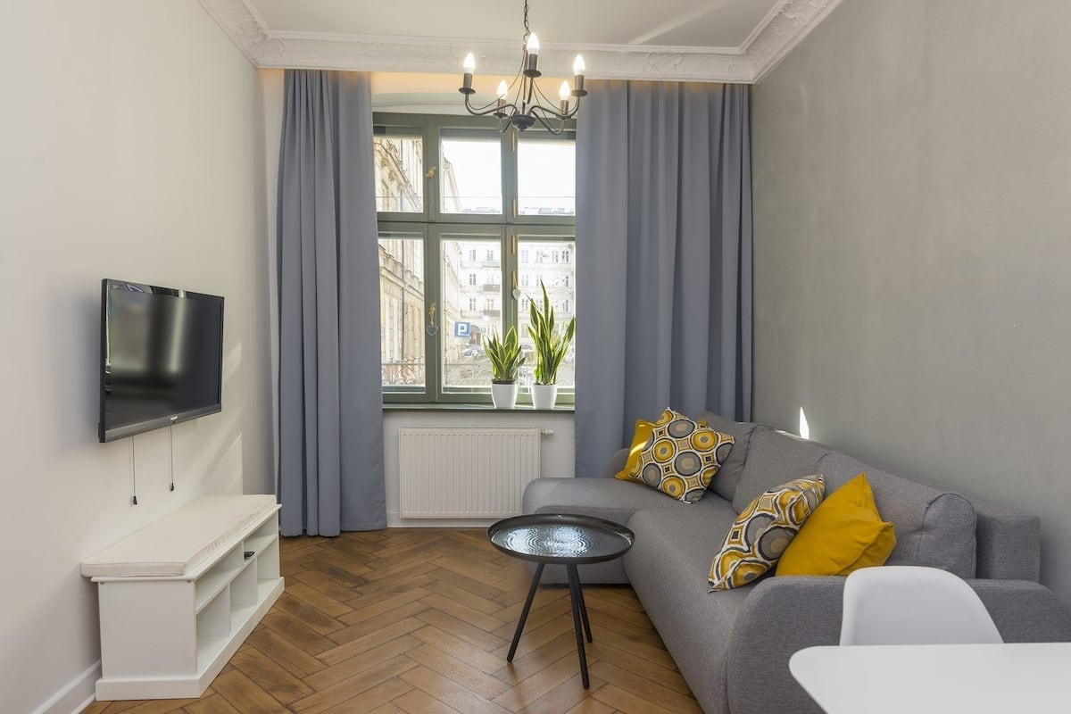 A modern living room with a flat-screen TV, grey sofa with patterned cushions, a small round table, and a window with curtains.
