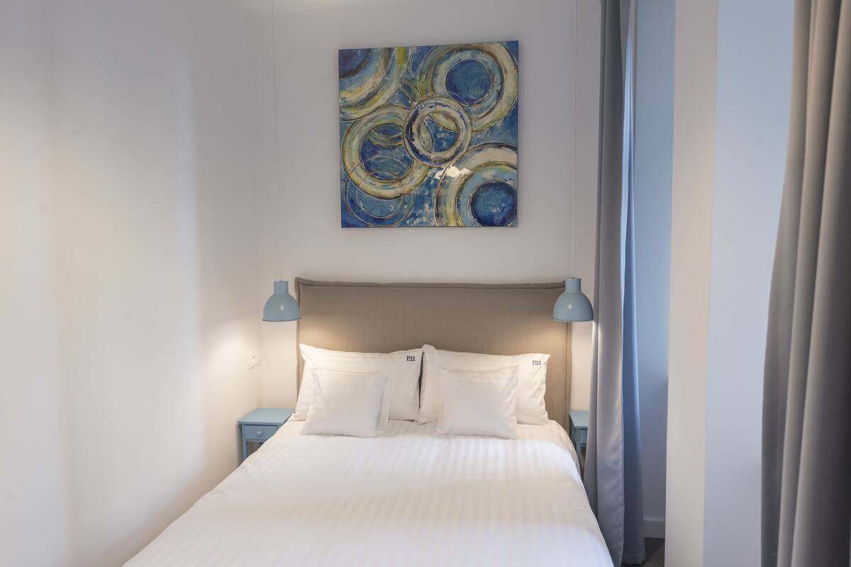 A neatly made bed with white linens, a blue abstract painting above it, and two bedside lamps on matching nightstands.