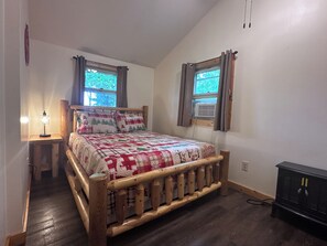 Een slaapkamer met een houten bed, twee ramen met gordijnen, een bedlampje en een klein kastje.