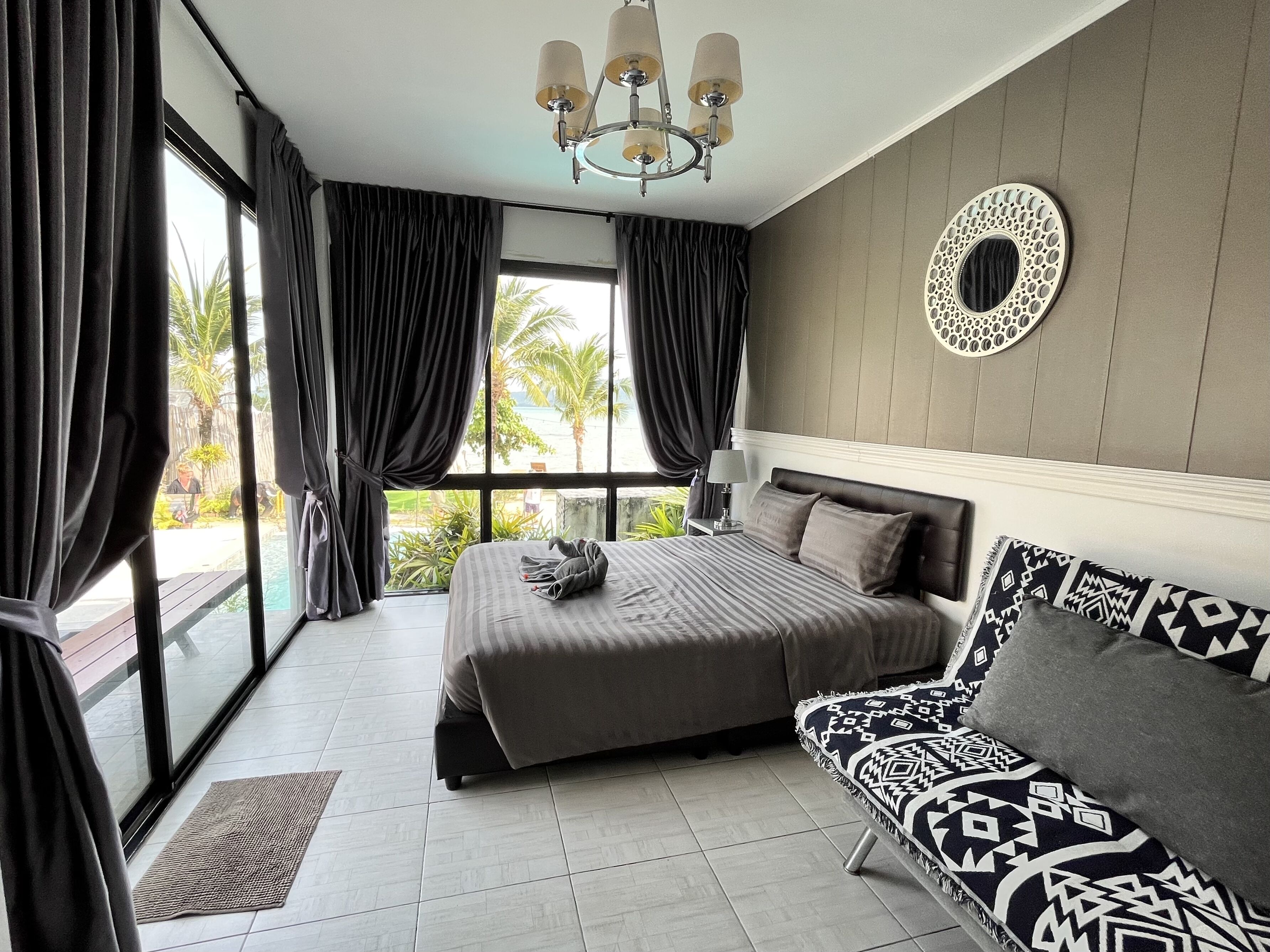 A bedroom with a bed, a sofa, a chandelier, a window with curtains, and a view of the outdoors.