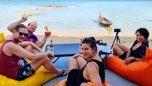 En gruppe mennesker slapper af i farverige pufposer ved en strand, med et kamera på stativ, en båd i vandet og et bjerg i baggrunden.