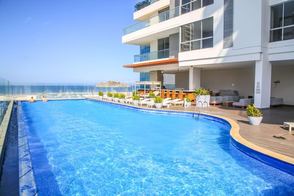 A modern hotel with a large swimming pool, outdoor seating, and a view of the ocean.