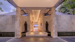 A long, narrow corridor with a stone floor, flanked by two large statues, leading to a bright interior space.