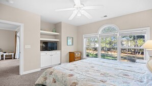 Un dormitorio con una cama grande, un ventilador de techo, un televisor sobre un aparador y una ventana con vistas a árboles.